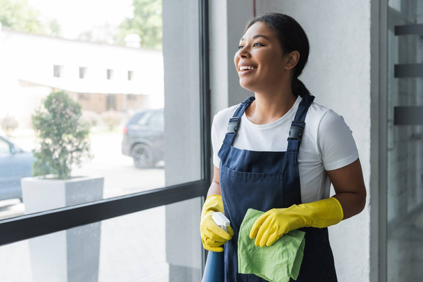 cheerful bi-racial woman with rag and detergent looking through window in office - Photo, Image