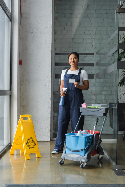happy bi-racial woman holding detergent and empty business card neat cart with cleaning supplies - Photo, Image