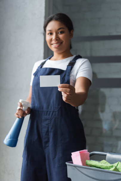 blurred bi-racial woman with spray bottle showing blank business card - Фото, изображение