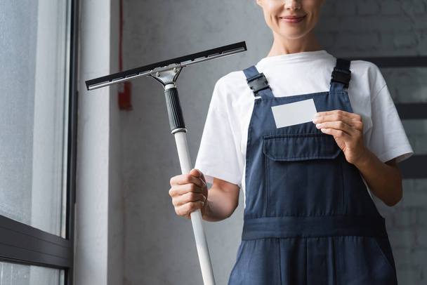 cropped view of smiling woman in workwear holding window squeegee and empty business card - Fotó, kép