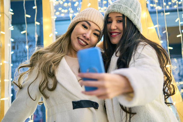Δύο κοπέλες ασιάτισσες με λευκά παλτά διασκεδάζουν αγκαλιασμένες και σέλφι στο κινητό, την Πρωτοχρονιά, λάμπες. - Φωτογραφία, εικόνα