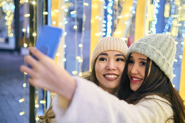 Dos amigas asiáticas con abrigos blancos se divierten abrazando y selfie en el teléfono móvil, año nuevo, bombillas - Foto, Imagen