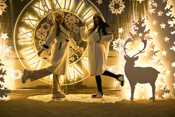 Δύο κοπέλες ασιάτισσες με λευκά παλτά διασκεδάζουν αγκαλιασμένες, νέα χρονιά, λάμπες. - Φωτογραφία, εικόνα