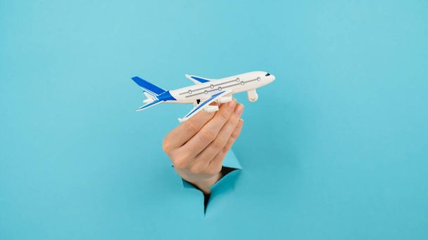 A female hand sticking out of a hole from a blue background holds a model of an airplane - Φωτογραφία, εικόνα