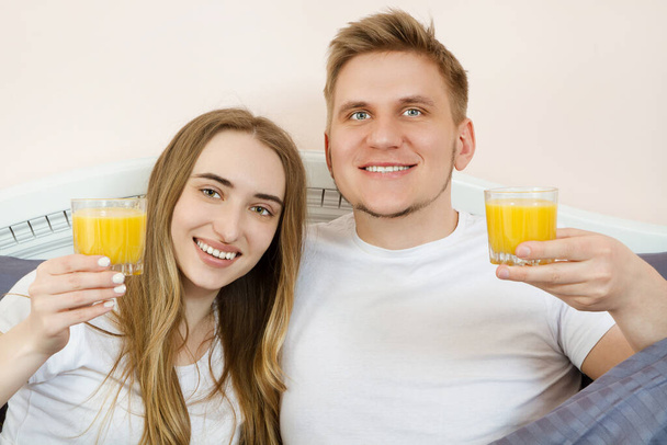 Happy relaxed young couple with orange juice in bed at home. Romantic couple drinking orange juice lying on their bed. - Foto, Imagem