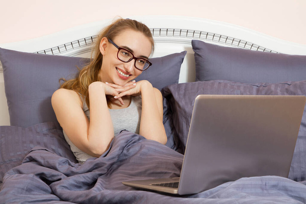 Hermosa chica en gafas se comunica a través de la computadora por chat de vídeo y se encuentra en la cama. Mujer feliz trabajando en el ordenador portátil en el dormitorio. - Foto, imagen
