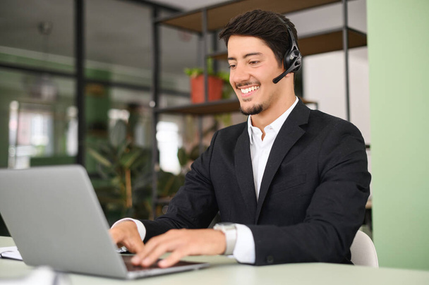 Cheerful male office employee in headset using laptop sitting on workplace, having video chat with employer or clients, business man using headphones with mic for online communication with customers - Photo, image