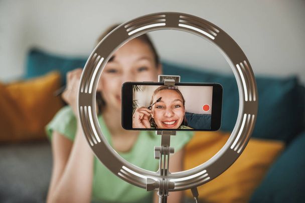 A cute teen girl vlogger live broadcasting make up tutorial to her followers via smart phone at her home. - Foto, Imagen