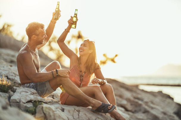 Young couple having fun and drinking beer on the beach by the sea. Enjoying in beautiful sunset and their love. - Fotoğraf, Görsel