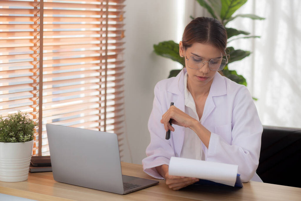Молодая женщина врач с помощью ноутбука компьютер и глядя документ о пациенте для диагностики в клинике, врач женщина рабочий и читающий документ в буфер обмена, один человек, медицинская концепция. - Фото, изображение