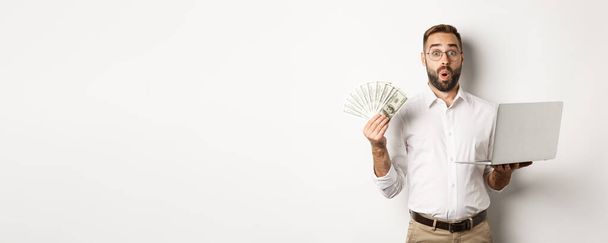 Business and e-commerce. Man looking amazed with money income, working online, using laptop, standing over white background. - Photo, Image