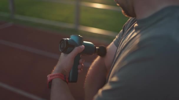 Athletic male massages muscles with hand massage gun, recovering from stadium running workout. . Body treatment with handheld wireless professional vibration shock massager after training session - Video