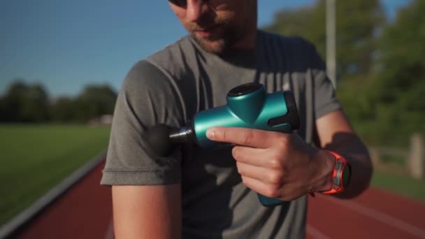 Athletic male massages muscles with hand massage gun, recovering from stadium running workout. . Body treatment with handheld wireless professional vibration shock massager after training session - Video