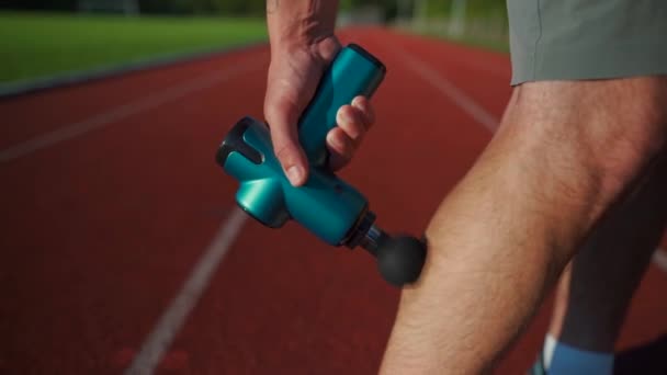 Athletic male massages muscles with hand massage gun, recovering from stadium running workout. . Body treatment with handheld wireless professional vibration shock massager after training session - Footage, Video