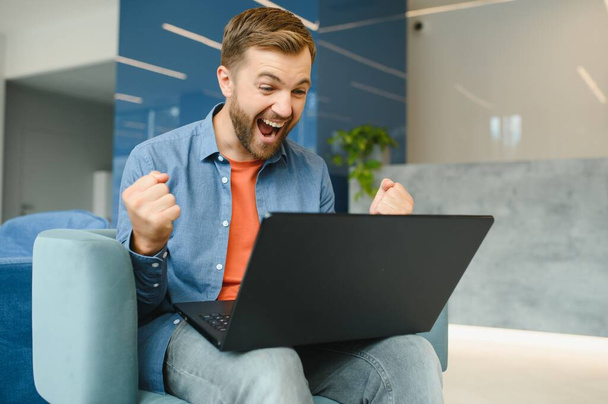 Photo of young excited man happy positive smile celebrate win victory success fists hands sit sofa home remote work - Photo, image