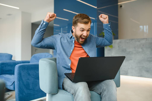 happy smiling remote online working man in casual outfit with laptop in joyful successful winning gesture sitting in an coworking office at a work desk - Φωτογραφία, εικόνα