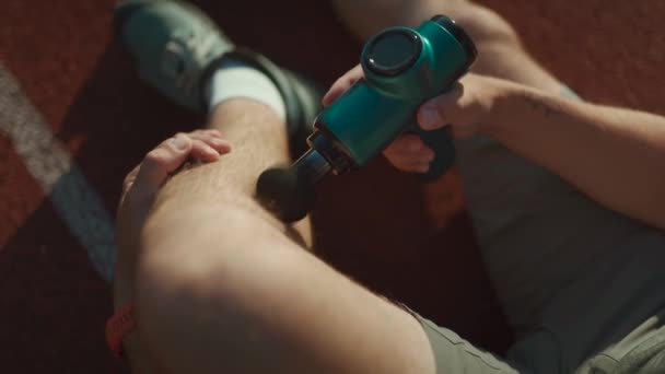 A male athlete massages muscles and tendons with a massage percussion device after a workout at the stadium. Sportsman uses electric massager gun in hand massaging the muscle. Sports recovery concept - Кадры, видео