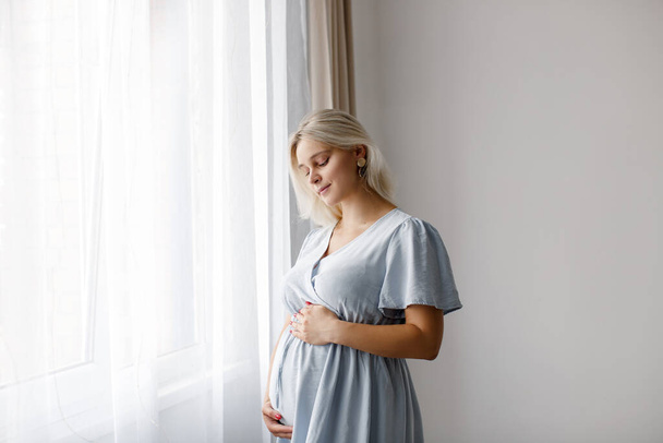Блондинка вагітна жінка в блакитній сукні стоїть біля вікна і торкається її живота. Концепція вагітності
.  - Фото, зображення