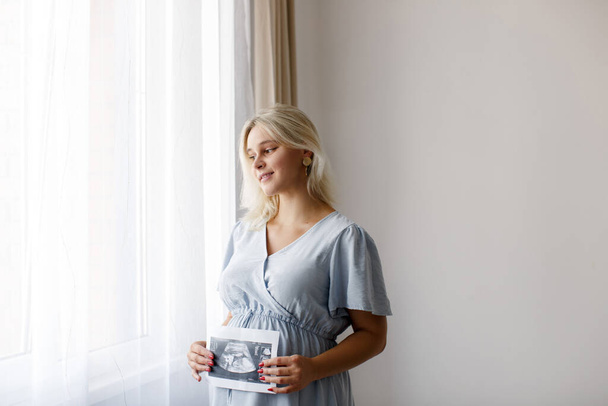A pregnant woman is holding ultrasound baby image near the window at home. The concept of pregnancy. - Foto, Bild