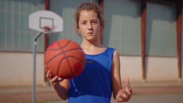 Joyful young sportsman posing with ball in sunlight on outdoor court. Portrait of smiling caucasian man standing at the background of basketball hoop and looking at camera. High quality 4k footage - Кадры, видео