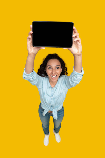 Happy Middle Eastern Lady Showing Digital Tablet With Empty Screen Advertising Website Posing Standing Over Yellow Studio Background. Vertical Shot, High Angle Shot - Foto, Imagen