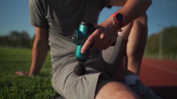 A male athlete massages muscles and tendons with a massage percussion device after a workout at the stadium. Sportsman uses electric massager gun in hand massaging the muscle. Sports recovery concept - Metraje, vídeo