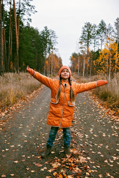 Happy ten years girl in orange coat with outstretched arms enjoys the fresh air in fall forest. Dry grass and golden trees. Autumn vibes, outdoor lifestyle. Emotional feeling, thinking and mental - Photo, image