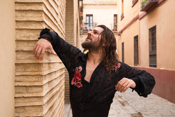 Long haired man dancing flamenco with black shirt and red roses. He makes dancing postures with his hands in a typical narrow street of Seville. Flamenco dance concept cultural heritage of humanity. - Фото, изображение