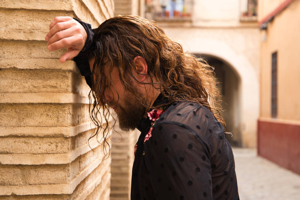 long-haired man dancing flamenco with black shirt and red roses rests his head on the wall of a narrow street typical of seville. Concept disappointment, depression, sadness, impotence, pain, passion. - 写真・画像