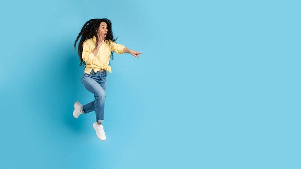 Shocked Middle Eastern Woman Pointing Finger Aside Jumping In Mid Air Showing Free Space For Text Posing Over Blue Studio Background. Panorama, Full Length Shot - Fotografie, Obrázek