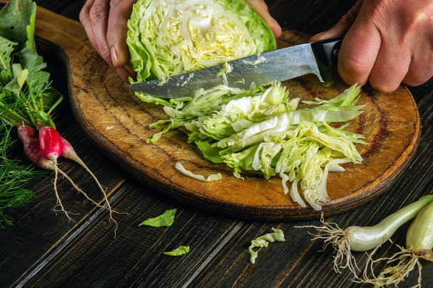 The cook cuts fresh cabbage with knife for salad on a kitchen table with fresh vegetables. Cooking and restaurant or hotel concept - Foto, Imagem