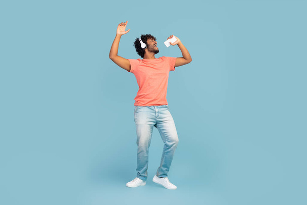Cool eastern millennial guy with curly hair singing and dancing on blue studio background, using mobile phone as microphone and wireless headphones, blue studio background, panorama, copy space - 写真・画像