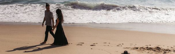男と女が手を取り合いながら海の近くのビーチでぬれた砂の上を歩く - 写真・画像