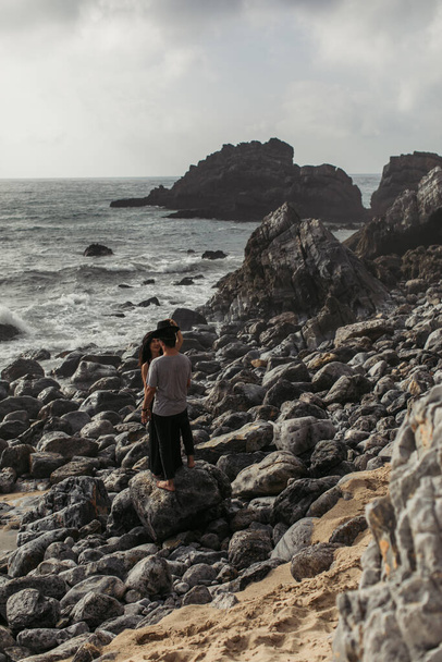 woman wearing hat on boyfriend while standing on rocks near ocean  - Photo, image