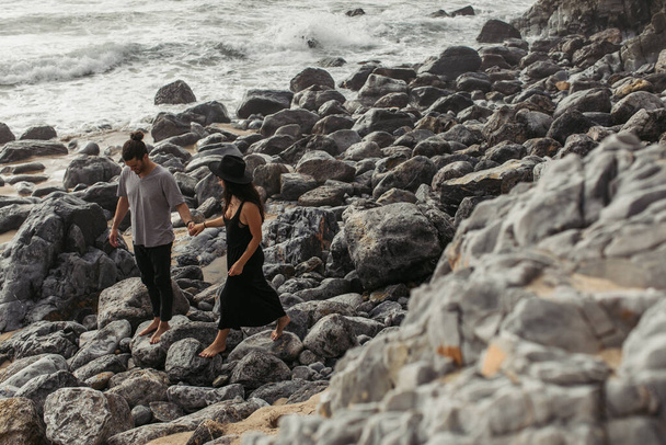 szakállas férfi mosolyog és kézen fogva barátnő ruha és kalap, miközben séta sziklák közelében óceán - Fotó, kép