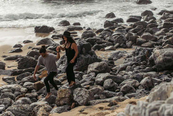 woman in hat holding hands with bearded man while walking on rocks near ocean - Photo, Image