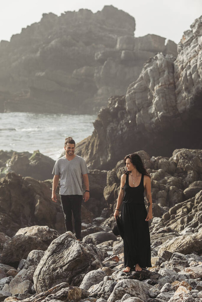 bearded man smiling while looking at tattooed girlfriend in dress while walking on rocks near ocean - Zdjęcie, obraz