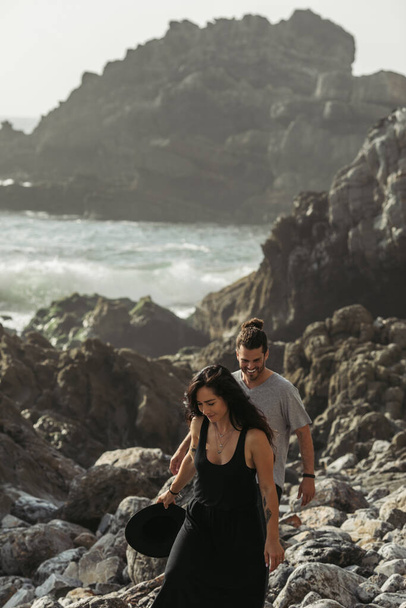 happy man smiling while walking on rocks near tattooed girlfriend in dress near ocean - Valokuva, kuva