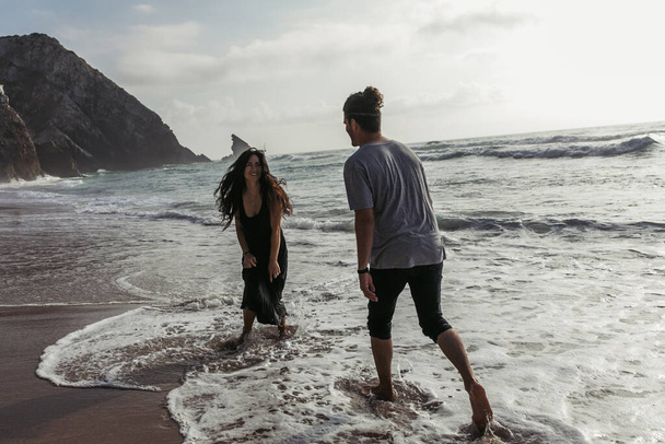 bearded man looking at happy girlfriend in dress standing in ocean water  - Photo, image