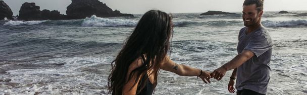 brodaty mężczyzna trzymający się za ręce z wytatuowaną dziewczyną w pobliżu oceanu, sztandar - Zdjęcie, obraz