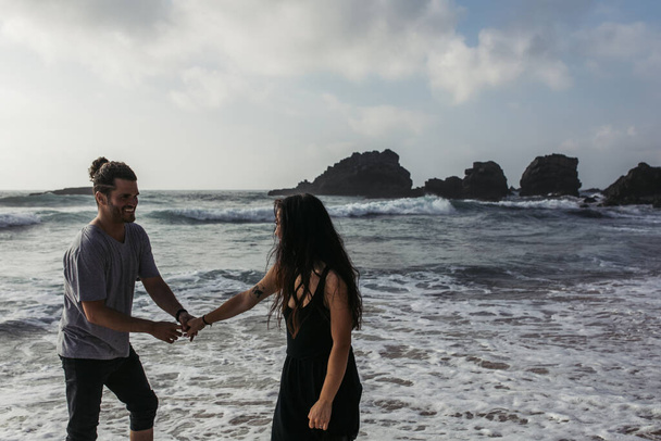 homem alegre de mãos dadas com a namorada tatuada no vestido enquanto estava perto do oceano  - Foto, Imagem
