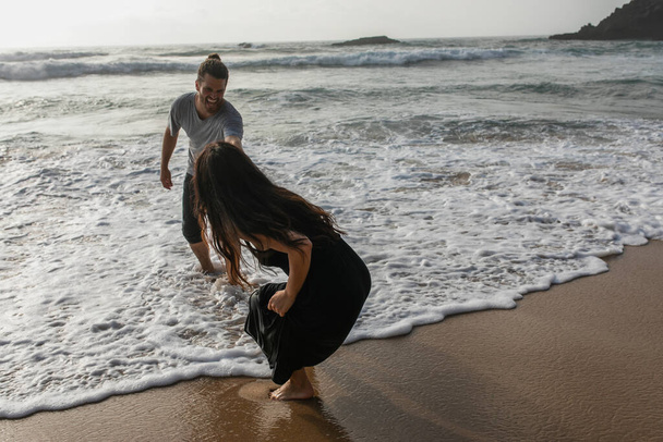 бородатый мужчина улыбается во время веселья с подругой в платье возле океана - Фото, изображение
