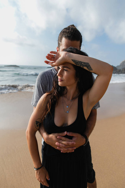 άντρας αγκαλιάζει κοπέλα με τατουάζ με φόρεμα κοντά στον ωκεανό κατά τη διάρκεια των διακοπών - Φωτογραφία, εικόνα