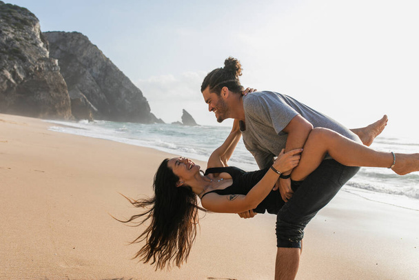 бородатый мужчина улыбается, поднимая женщину в платье и веселясь на пляже возле океана - Фото, изображение