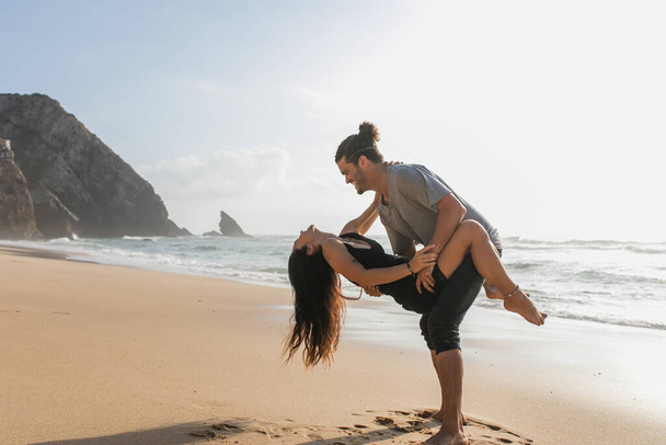 side view of happy bearded man lifting tattooed woman in dress on beach near ocean  - Photo, image