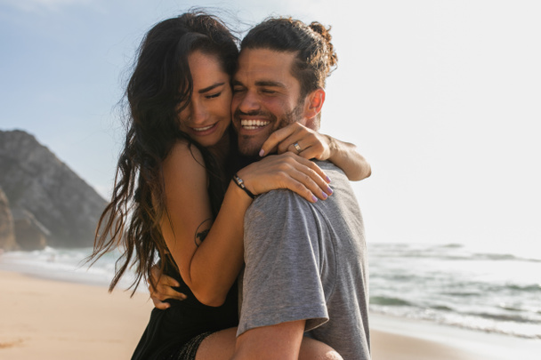cheerful man lifting and hugging tattooed woman in dress on beach near ocean  - Photo, image
