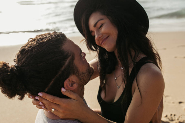 щаслива жінка в капелюсі посміхається, дивлячись на бородатого хлопця на пляжі
 - Фото, зображення