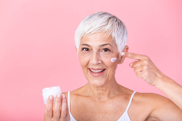 Beautiful mature woman holding jar of moisturizer and looking at camera. Happy senior woman holding a bottle of anti-aging lotion isolated over pink background. Anti aging and beauty treatment. - Foto, Imagen
