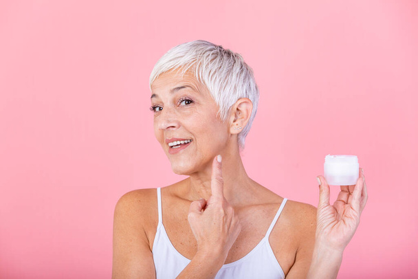 Beautiful mature woman holding jar of moisturizer and looking at camera. Happy senior woman holding a bottle of anti-aging lotion isolated over pink background. Anti aging and beauty treatment. - Foto, imagen