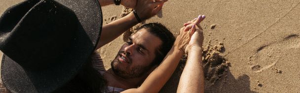 vista superior del hombre barbudo cogido de la mano con la mujer en el sombrero y acostado en la arena, bandera - Foto, Imagen